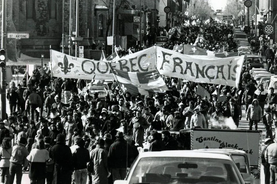 Une foule défile rue Saint-Antoine, à Montréal, derrière une banderole « Québec français » et des drapeaux du Québec